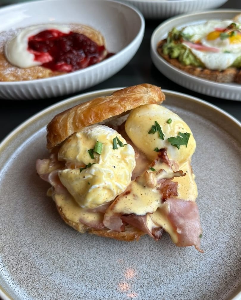 Croissant Eggs Benedict with ham, hollandaise, and chives, alongside other breakfast dishes.