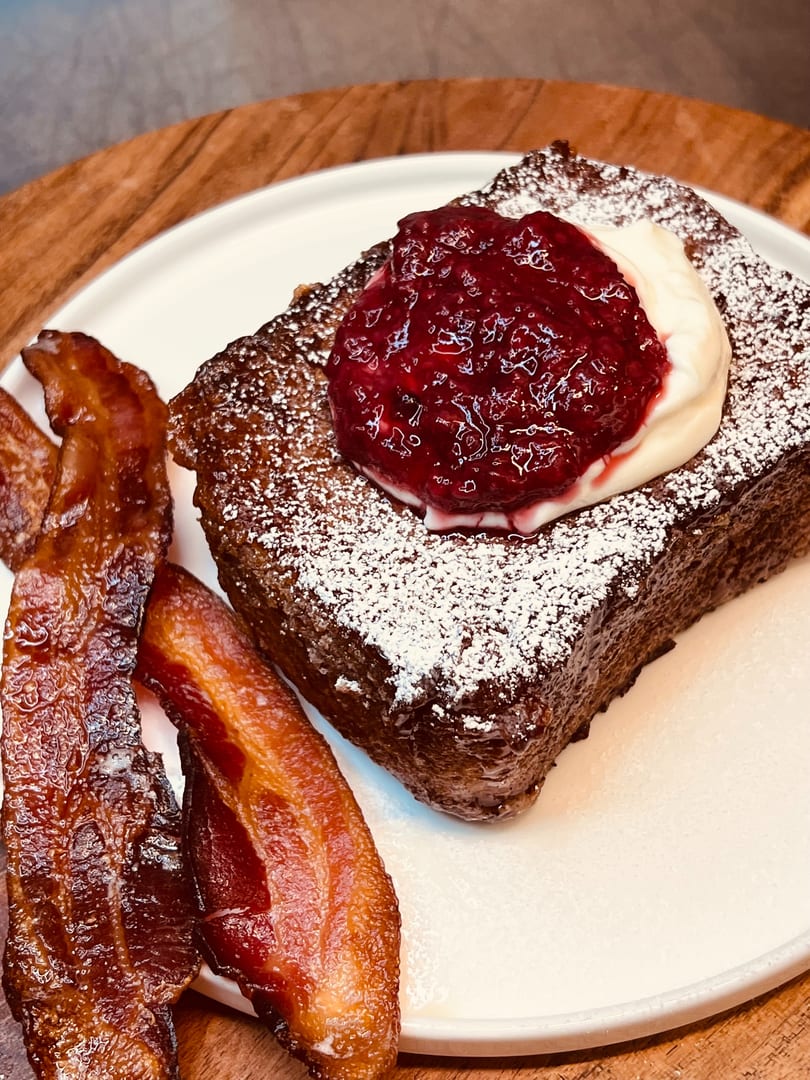 Thick French toast with powdered sugar, berry compote, whipped cream, and crispy bacon on a white plate.