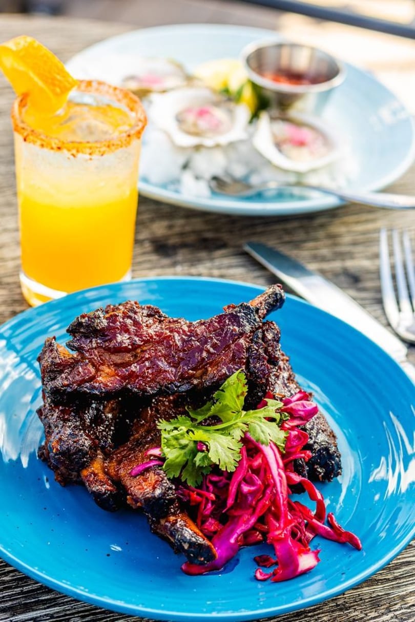 Glazed ribs with red cabbage slaw and cilantro on a blue plate, with a drink and oysters.