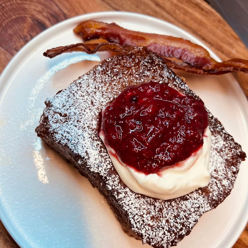 Thick-cut French toast with powdered sugar, whipped cream, berry compote, and a side of bacon.