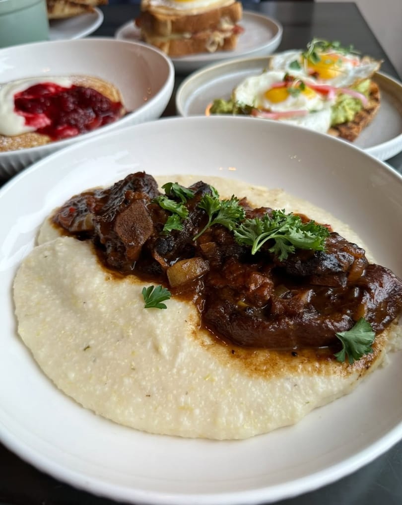Braised short ribs with creamy grits, garnished with parsley, alongside other breakfast dishes.